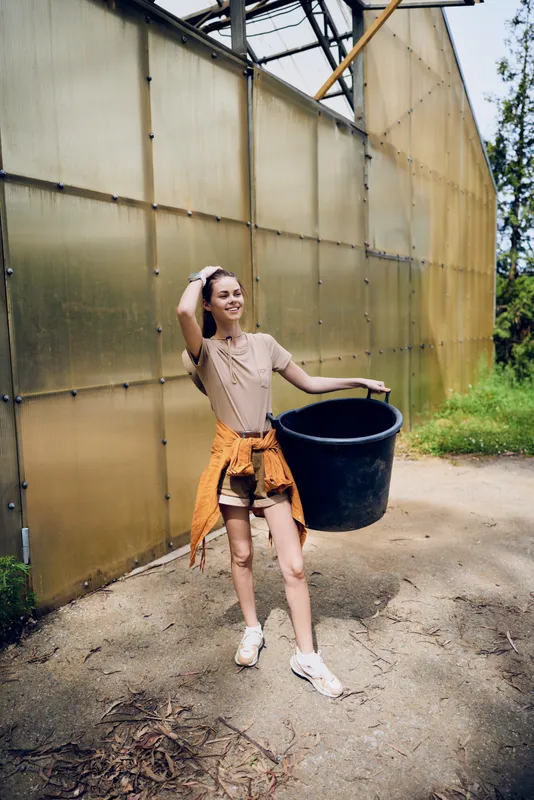 Woman young smiling while gardening outdoors, carrying a bucket near a greenhouse in casual clothes. Full body portrait of female doing farm work with container, summer lifestyle and care.