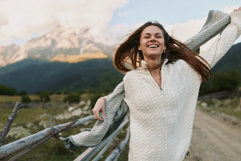 Happy woman in white sweater enjoying fresh air outdoors near wooden fence with mountains in background. Concept of lifestyle, freedom, and nature relaxation.