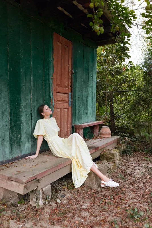 Woman sits on a wooden bench outside a green rustic house wearing a pale yellow dress, relaxed and smiling, enjoying sunlight, greenery, and a peaceful countryside scene