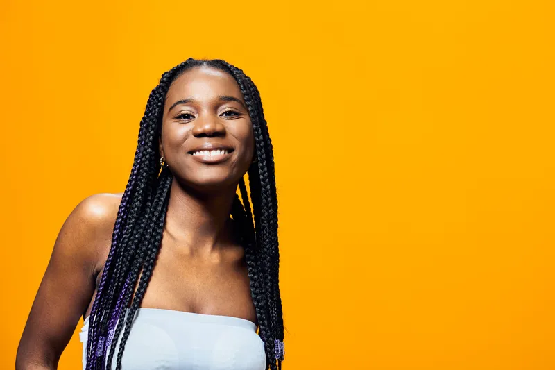 Smiling Black woman with braided hair on a vibrant yellow background, showcasing confidence and joy in a casual outfit, ideal for lifestyle and wellness concepts