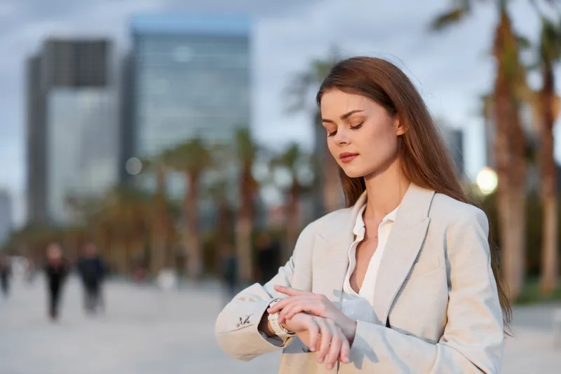 Woman in business attire checking wristwatch outdoors with modern city skyline and palm trees in background, focus on time management and professionalism