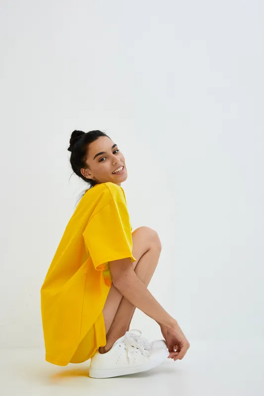 Young woman wearing oversized yellow t-shirt and white sneakers crouching on white background, smiling and relaxed with casual style and simple composition.