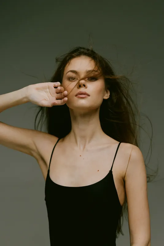 Young woman with flowing hair posing in black dress with neutral gray background, elegant and minimalistic style