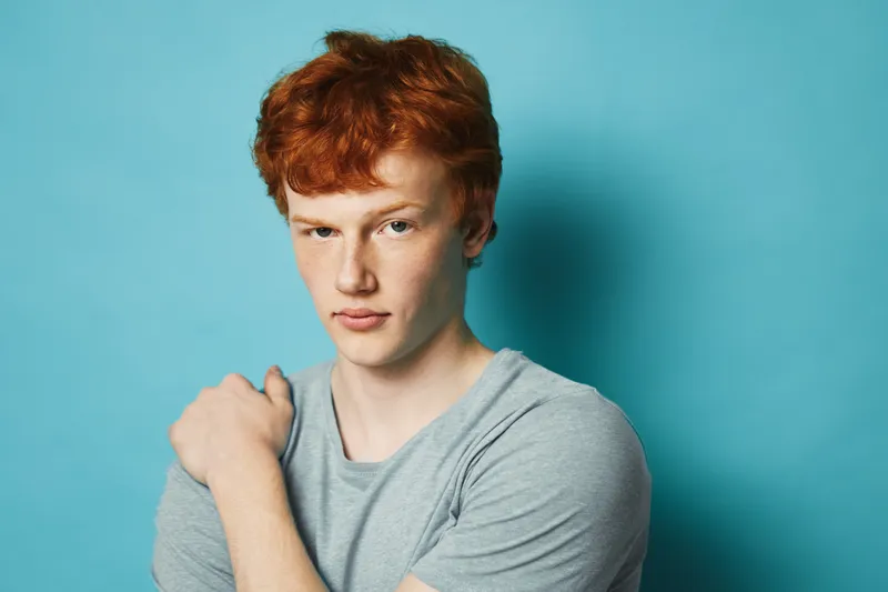Young man with red hair wearing gray shirt posing with arm crossed against blue background. Portrait of confident male model with casual style and serious expression.