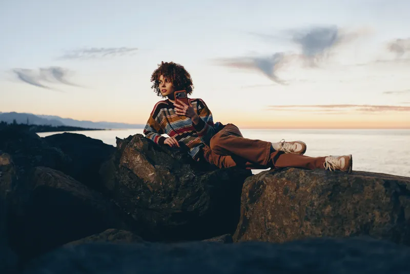 Lifestyle woman sitting on cliff rocks at sunset, striped top and casual shorts, relaxed pose with legs on stone, ocean horizon and distant hills behind, warm calm mood, outdoor golden hour