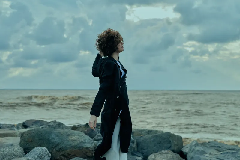 Woman in a dark coat stands on a rocky shore beside the sea, wind blowing, cloudy sky, capturing moody outdoor coastal atmosphere