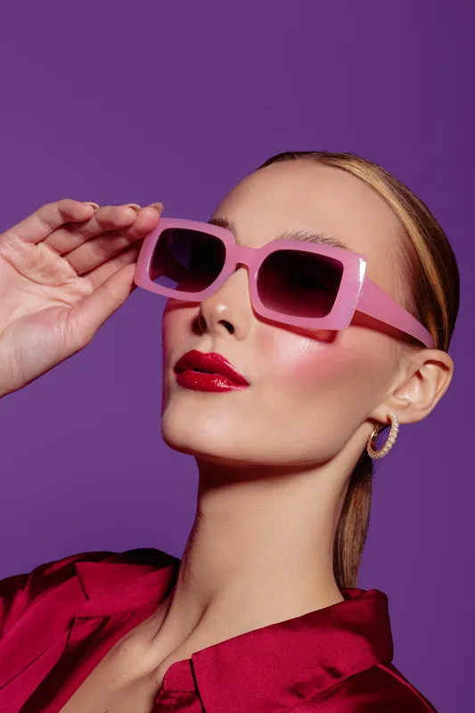 Stylish woman with pink sunglasses and red lipstick poses against purple background. Fashion portrait with glossy makeup and elegant hairstyle. Concept of beauty and modern style.