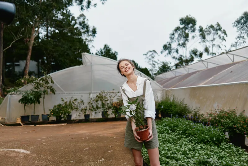 Happy young woman holding a potted plant in a greenhouse, wearing a stylish outfit, surrounded by greenery in a bright, natural setting