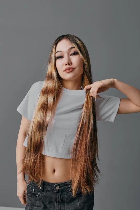 Young woman with long hair wearing cropped white t shirt and jeans posing against grey background looking confident and stylish indoors with natural light.