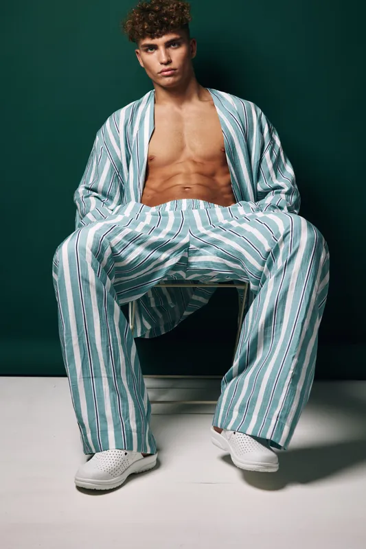 Young man with curly hair wearing striped loose pants and open shirt sitting on chair with serious expression in studio with dark green background.