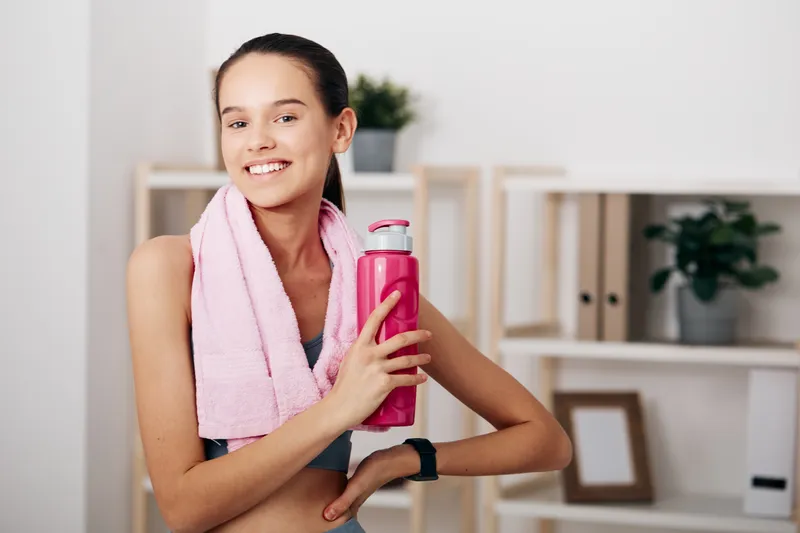 Fitness woman smiling after workout, holding a pink water bottle with towel draped over shoulder in a bright indoor gym, showcasing health, wellness and an energetic training vibe.