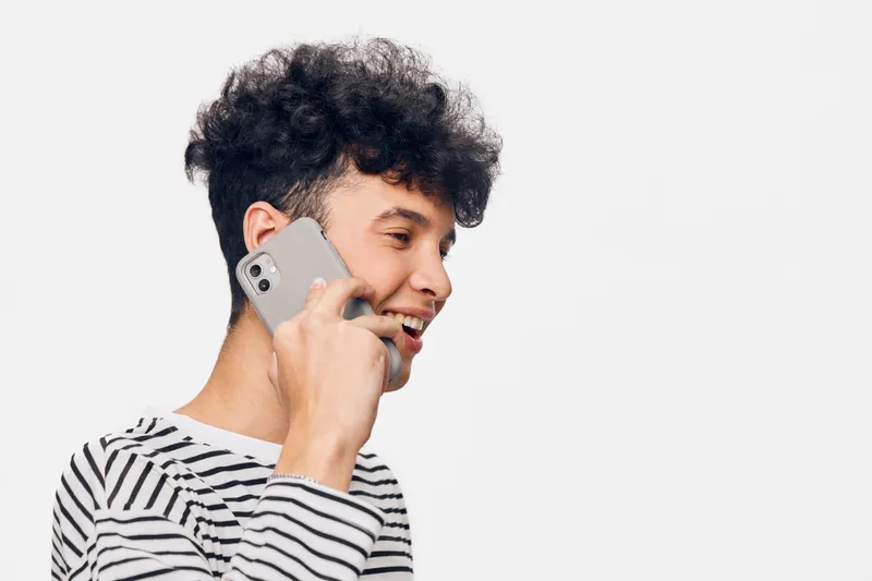 young man talking on smartphone wearing striped shirt, curly hair and smiling against white background in bright casual style