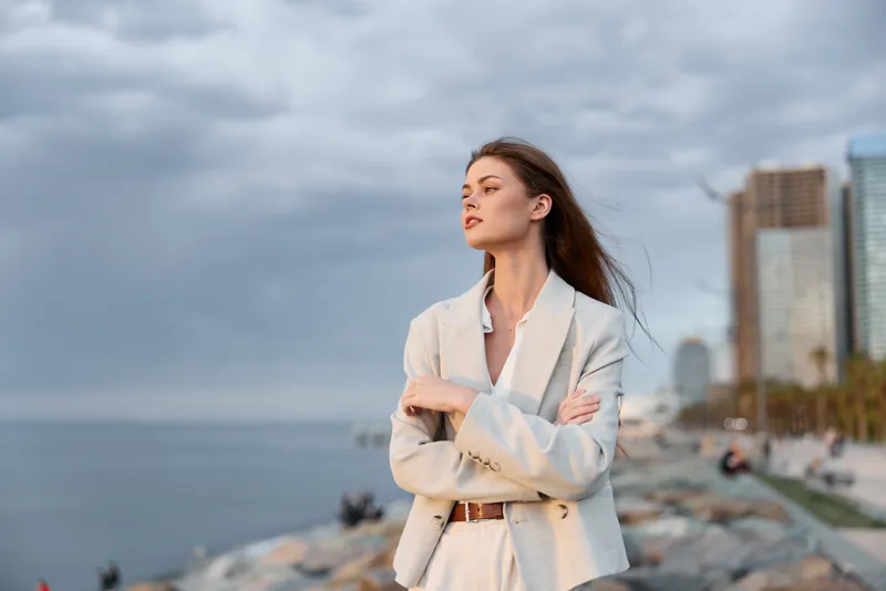 confident business woman in light suit standing with crossed arms near sea shore with city skyline and cloudy sky background