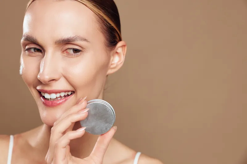 A young woman smiles while applying makeup with a powder compact in a soft studio light, a close-up portrait conveying a calm, everyday beauty moment.