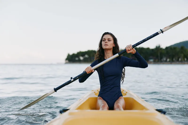 A young woman rows solo in a yellow rowboat on calm water, wearing a dark swimsuit, focused as she glides with the oar in hand.