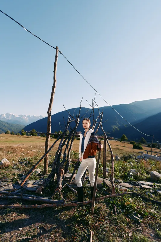 Man in traditional jacket stands by rustic fence in a rural field with mountains and wide landscape, portrait of countryside life, boots, clear sky and natural scenery.