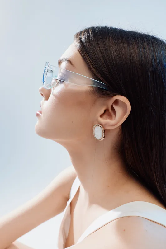Profile of a young woman wearing transparent glasses and elegant earrings. Soft lighting highlights her smooth skin and long dark hair. Concept of beauty fashion and style.