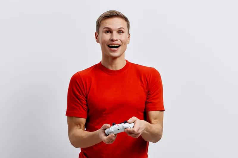 Vibrant young man in red t-shirt playing video game with white controller, smiling with excitement, isolated on plain light gray background, studio portrait. People lifestyle entertainment concept