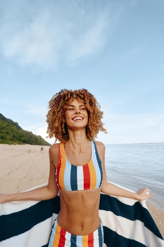 A joyful woman in a striped bikini stands on a sunny beach, holding a towel and smiling with open arms. The scene conveys carefree summer wellness, outdoor recreation, and travel spirit.