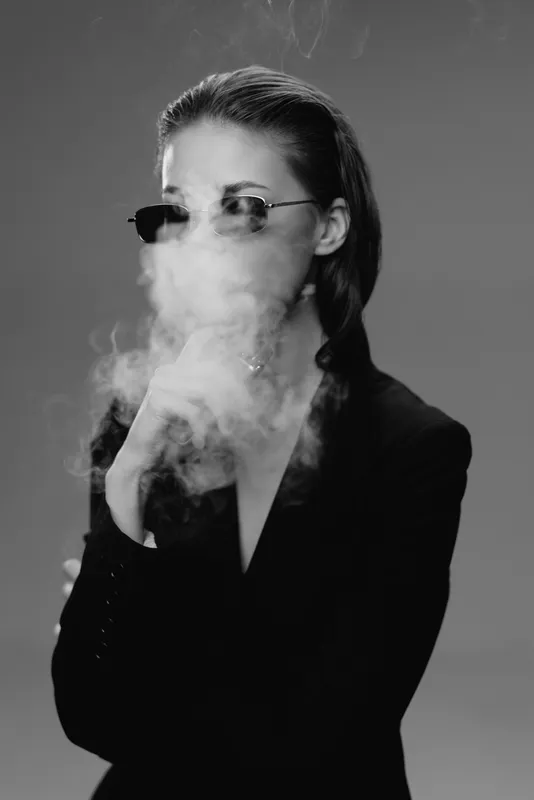 Stylish woman smoking with sunglasses in black and white, emitting smoke and wearing a black jacket.