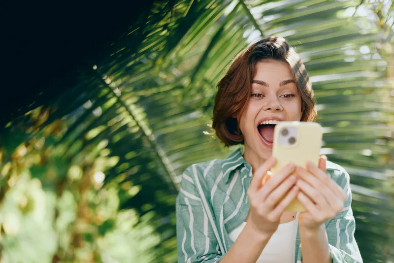 woman smiling selfie outdoors on a sunny day, casual striped shirt, handheld smartphone, tropical greenery in the background, bright natural light, joyful mood, candid portrait