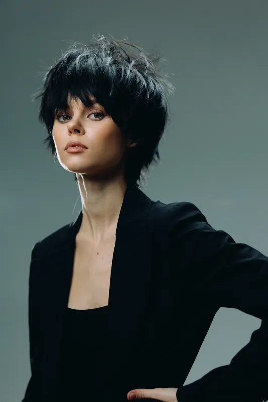 Stylish woman with short dark hair wearing black outfit posing against gray background in studio setting