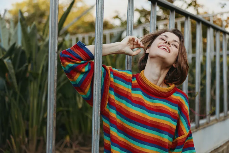 Woman in a colorful striped sweater smiling and leaning against a metal fence, short hair bob in an outdoor portrait among lush greenery, relaxed joyful mood for lifestyle and fashion travel.