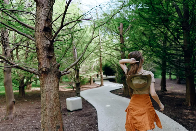Woman walking on winding path in a shaded forest, wearing a straw hat and orange skirt, back view on a tree-lined trail, serene nature scene and outdoor leisure escape.
