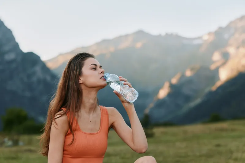A hydrated woman drinks water from a bottle in a scenic mountain landscape. She wears a sleeveless top and looks relaxed, enjoying the fresh air, sunlight, and tranquil outdoor surroundings.