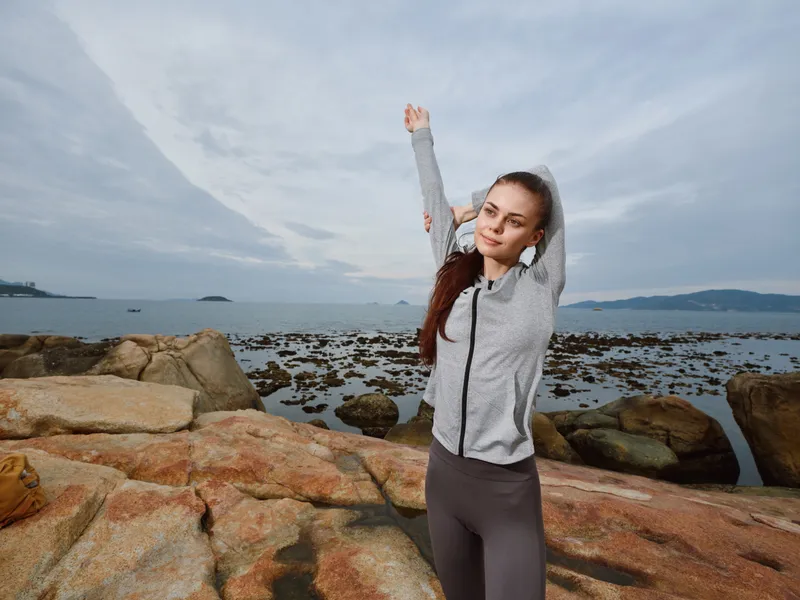 Young fitness model stretching outdoors by the sea wearing a gray jacket and black leggings, embodying healthy lifestyle and wellness concepts