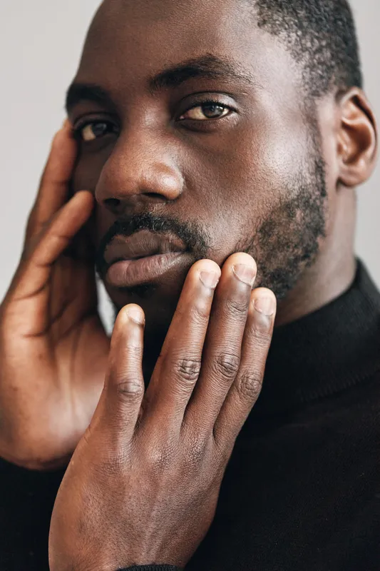 African man with dark skin and beard in creative conceptual visual style touching face with hands closeup portrait. Male model with thoughtful expression wearing black turtleneck sweater indoors.