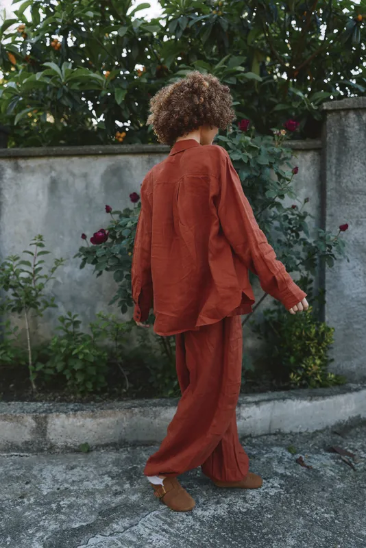 A woman in a rust colored suit walks along a garden wall, back turned, casual street style in an outdoor setting, emphasizing contemporary fashion and relaxed lifestyle