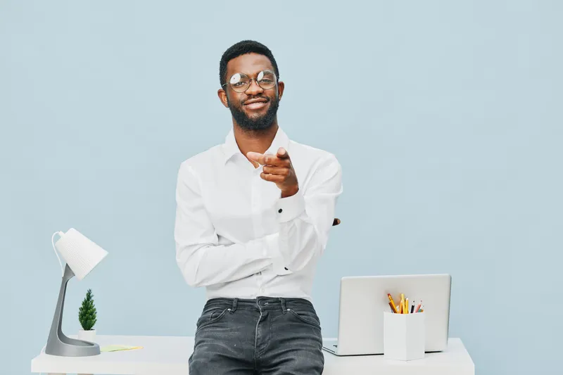 Confident smiling man wearing white shirt and glasses standing at modern office desk with laptop, desk lamp, and plant against light blue background. People lifestyle concept, professional workspace, 