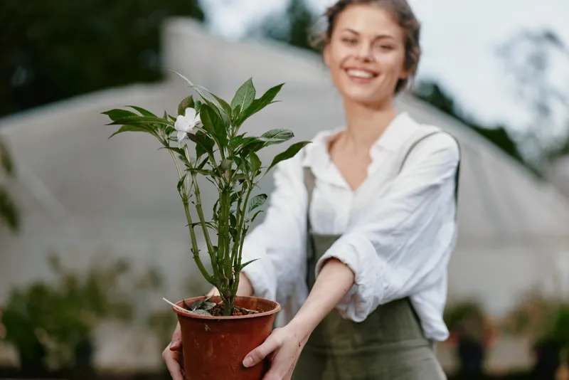 Cheerful young woman in a garden holding a potted plant, showcasing her passion for gardening and nature, with a soft focus background and natural light