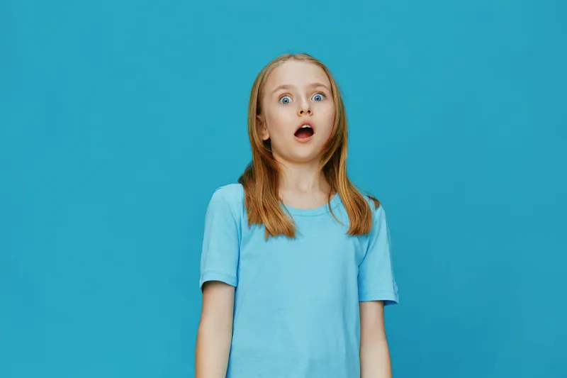 Surprised young girl with long hair in a blue t shirt against a bright blue background, expressing shock and wonder, evoking childhood emotions of amazement and curiosity