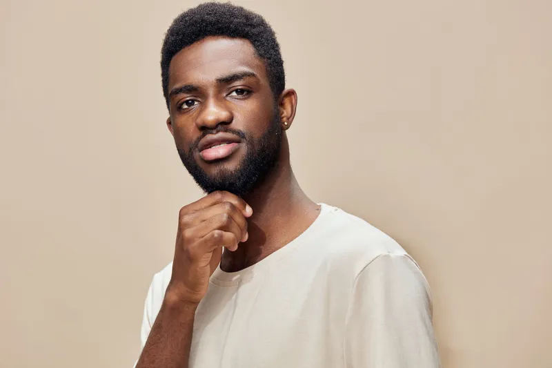 Young Black man with a confident expression, wearing a light colored shirt, posing against a warm beige background, ideal for lifestyle and fashion themes