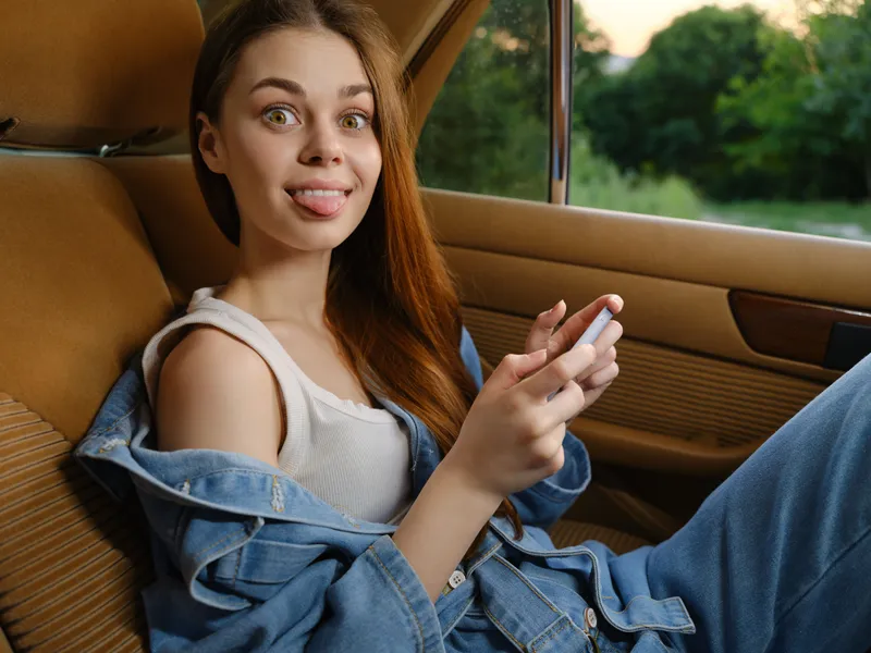 Happy young woman in casual outfit sitting in car with phone, joyful expression, outdoor background, summer vibes, denim style, vibrant colors.