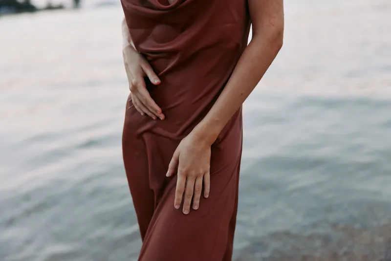 Close-up of a person in a deep burgundy dress by calm water, highlighting flowing fabric and a poised hand, capturing subtle elegance and soft, reflective light.