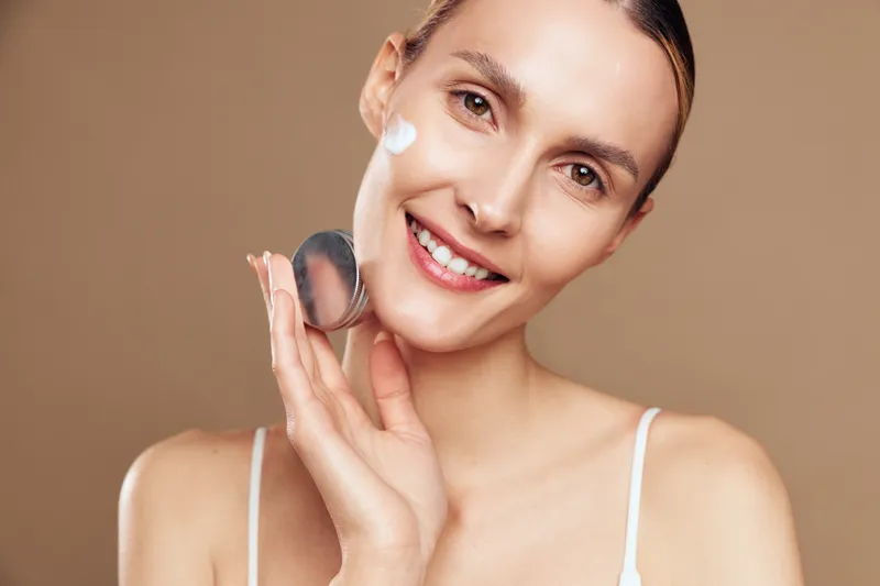 Woman skincare face beauty cream smile application, isolated portrait on neutral background, young blonde woman applying lotion, fresh natural glow, personal care