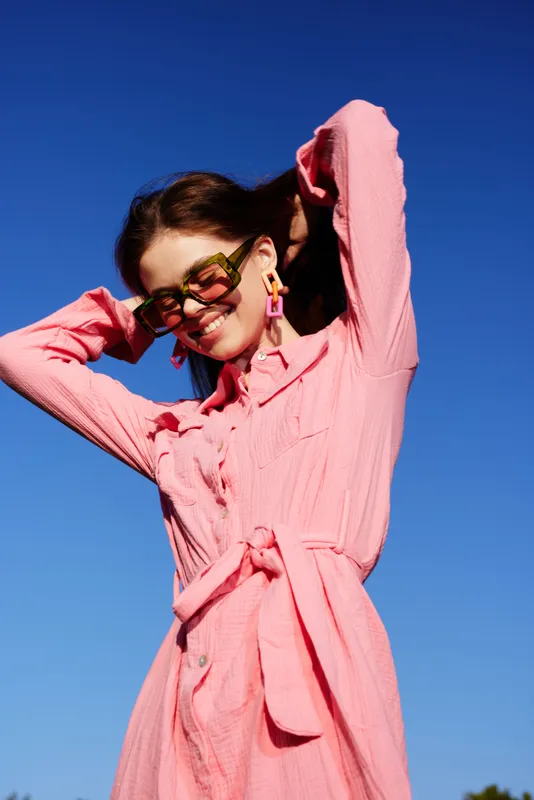 Cheerful young woman in a pink dress enjoying a sunny day, showcasing summer fashion and vibrant emotions against a clear blue sky