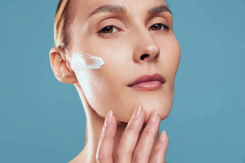A young woman applies moisturizing cream to her cheek, demonstrating skincare routine on her smooth face against a blue background. Concept for beauty, health, and self-care.