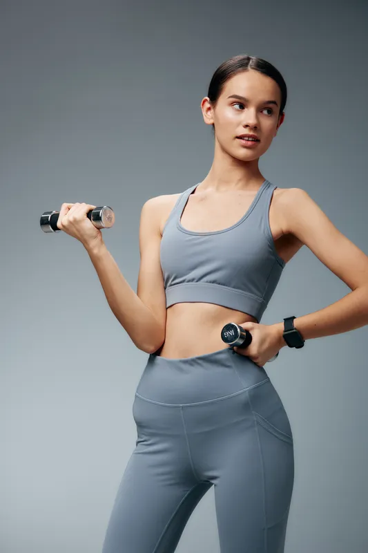 Woman in athletic gray sportwear holds dumbbells in a studio, showcasing fitness training and workout technique for health and athletic performance.