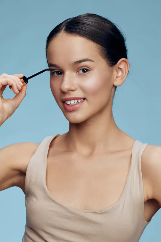 Young woman applying mascara, showcasing natural beauty and confidence against a soft blue background Ideal for beauty and makeup concepts