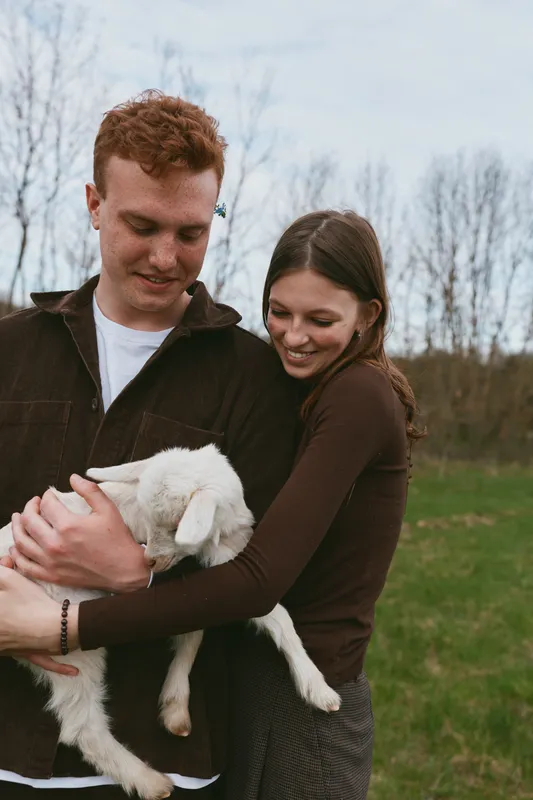 A couple outdoors warmly embraces a gentle goat kid in a grassy field, soft light, casual clothing. A sweet moment of connection, bonding, and rural lifestyle with animals.