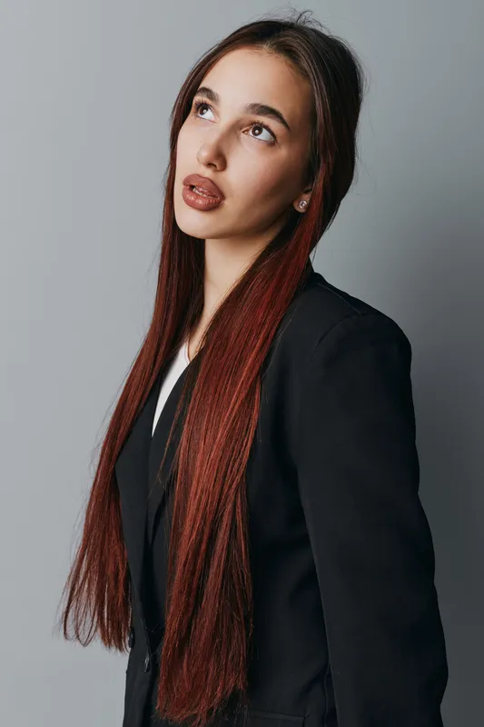 Young woman with long straight hair wearing black blazer looks upward with thoughtful expression on gray background. Portrait of confident female model with natural makeup and casual style.