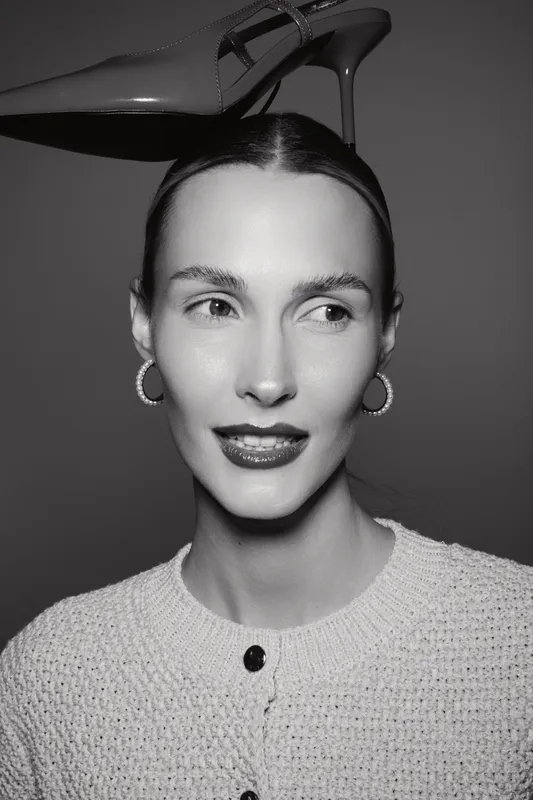 Portrait of a slim white woman with pale skin wearing hoop earrings and a textured cardigan. She balances a high heel shoe on her head against a dark background. Concept of fashion and elegance.