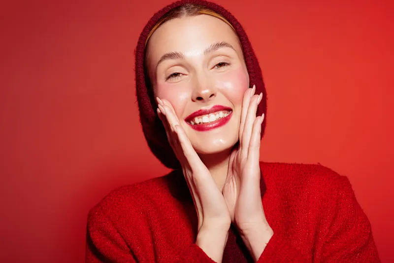 Smiling woman in red sweater and hat touches her face with hands on red background. Concept of happiness, beauty, fashion, and winter style with bright makeup and joyful expression.