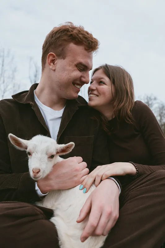 Young couple shares a tender moment outdoors as the man gently holds a white baby goat while they smile and look at each other with affection in a natural setting.