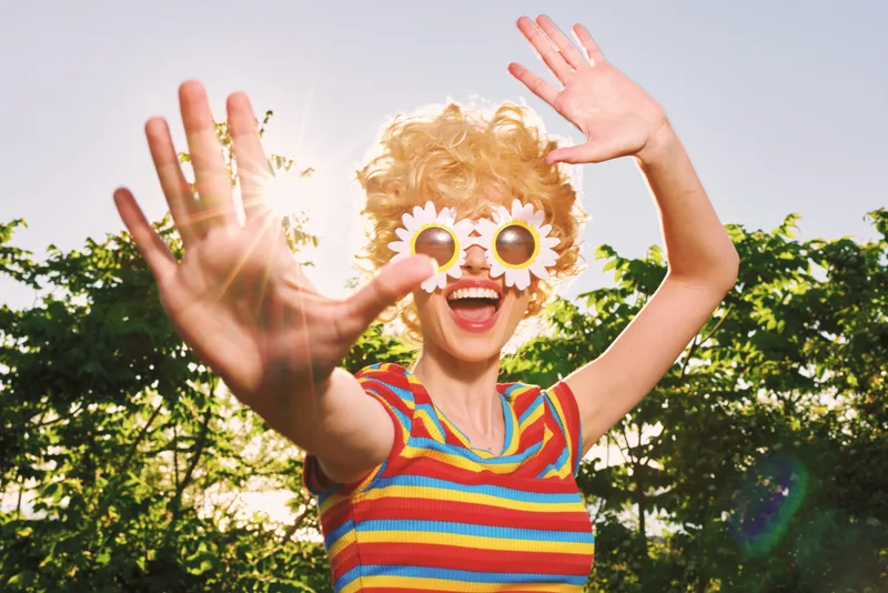 Playful woman outdoors wearing quirky glasses, smiling at the camera. Bright striped shirt, sunny day, retro color palette, candid lifestyle moment capturing carefree joy.