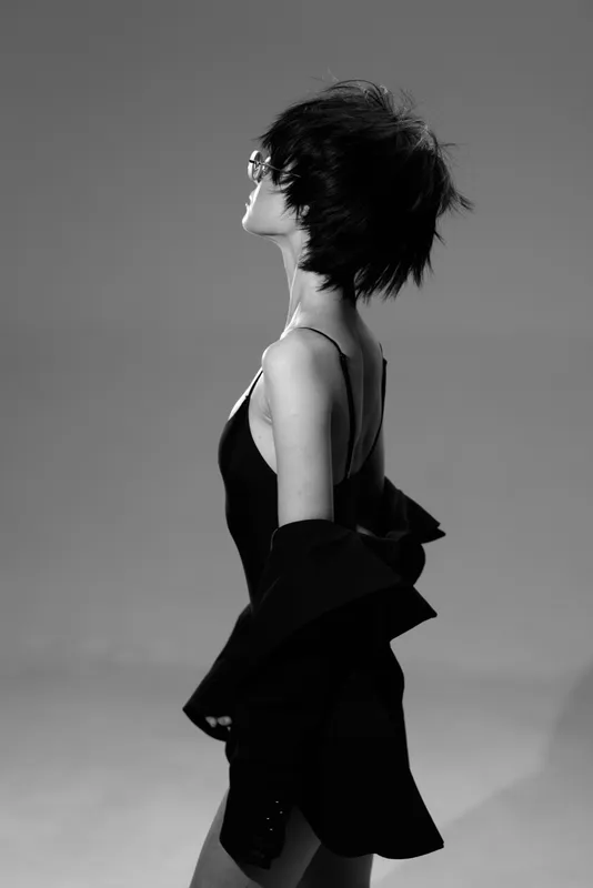 Black and white portrait of a stylish woman with short hair posing confidently in elegant clothing with soft lighting and minimalistic background.